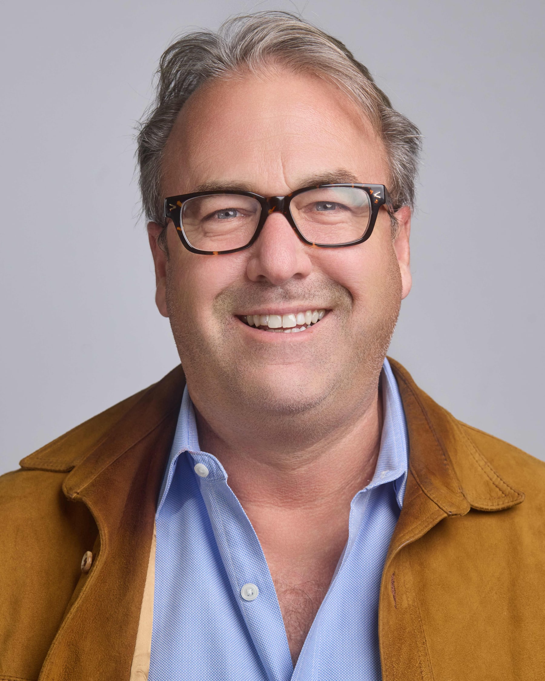 Studio headshot of a man in glasses and a tan jacket